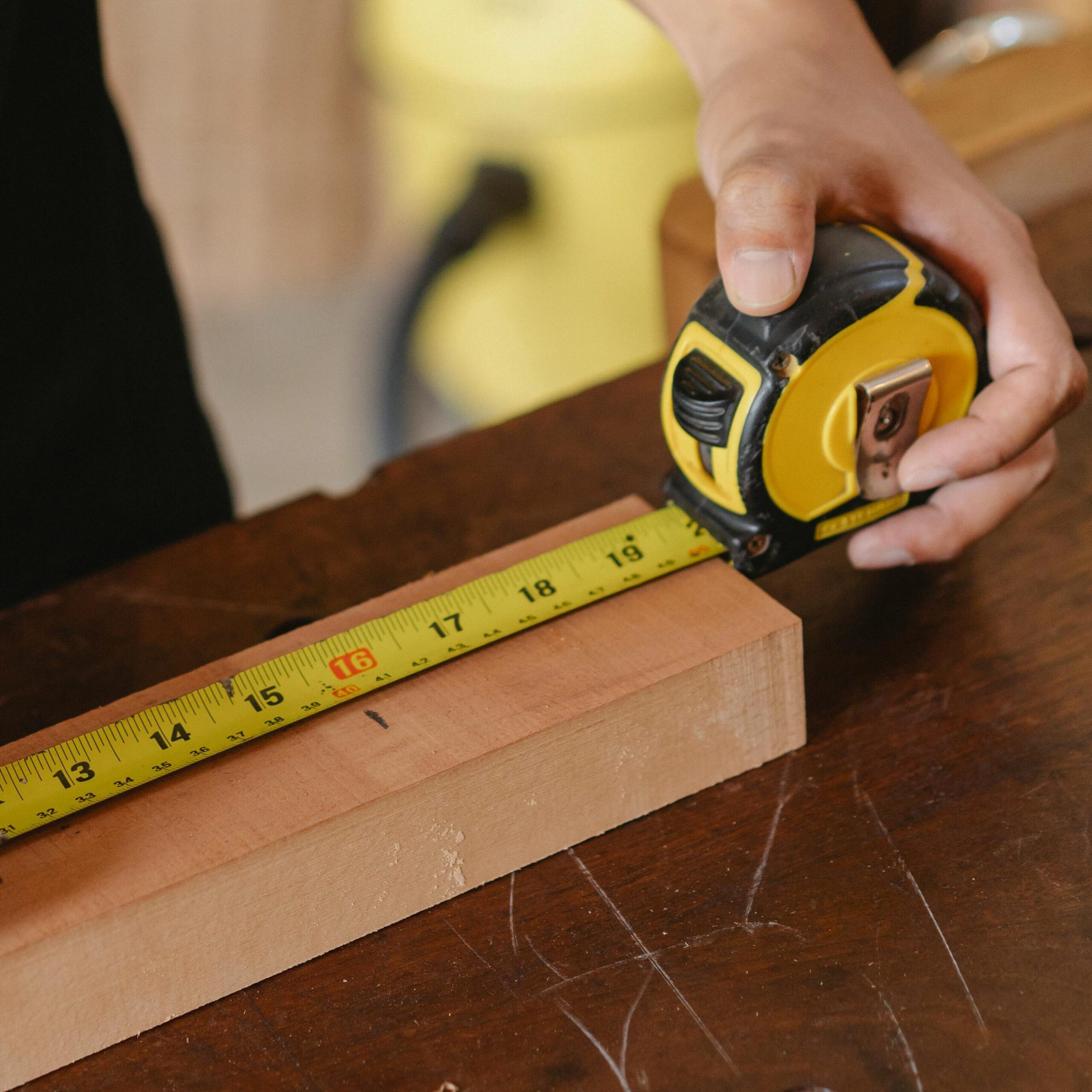 Accurate unrecognizable male worker with pencil and measuring tape on wooden board standing near workbench during work process on blurred background